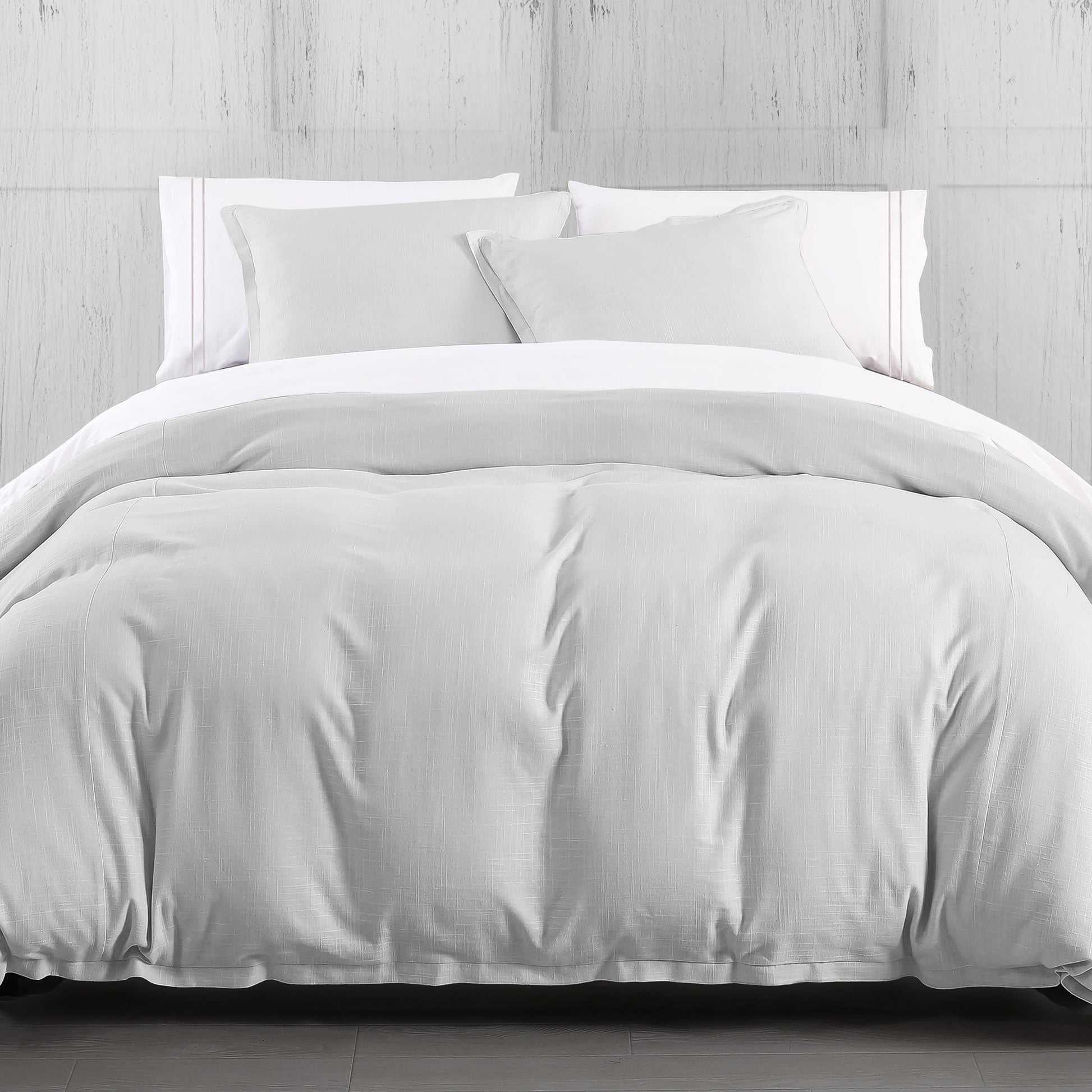 White comforter set on a bed with white pillows against a light wooden wall.