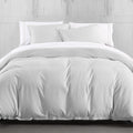 White comforter set on a bed with white pillows against a light wooden wall.