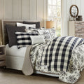 Bedroom with floral and checkered bedding, pillows, and a lamp.