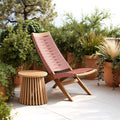 Pink outdoor lounge chair with wooden legs and a matching zahara side table natural on a patio with plants.